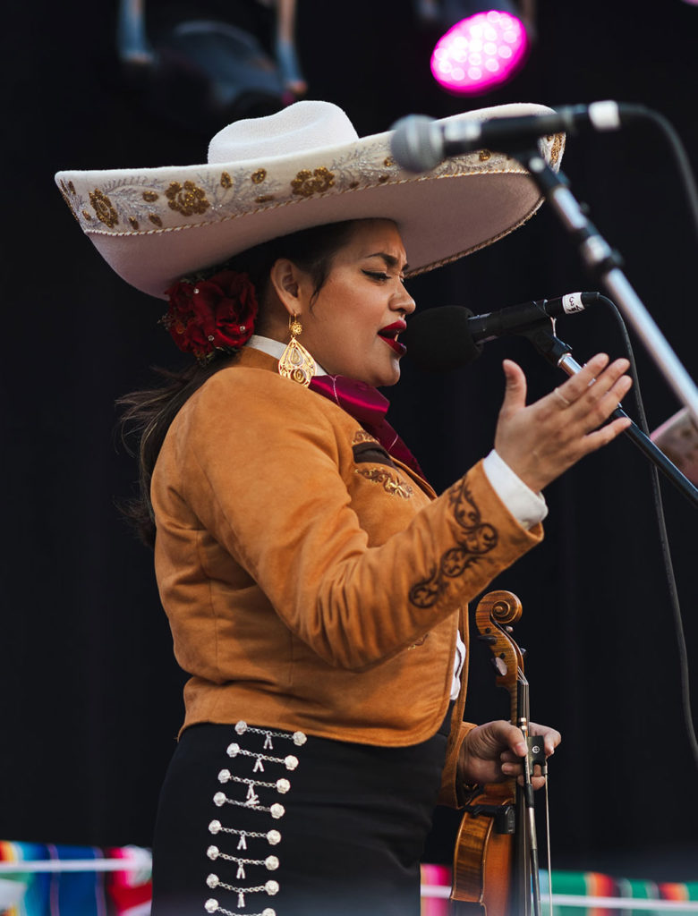 Woman wearing a large sombrero and mariachi uniform, singing and holding a violin