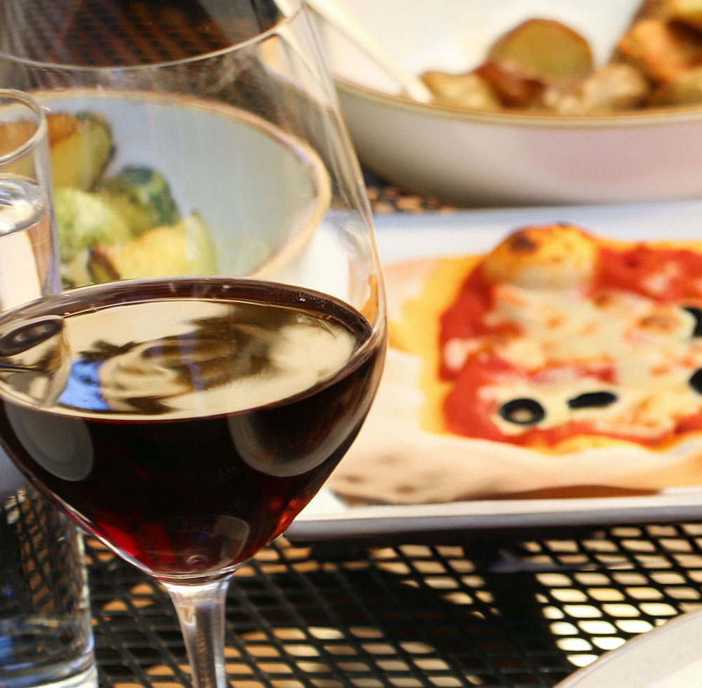 Photo of a glass of red wine with food in the background.