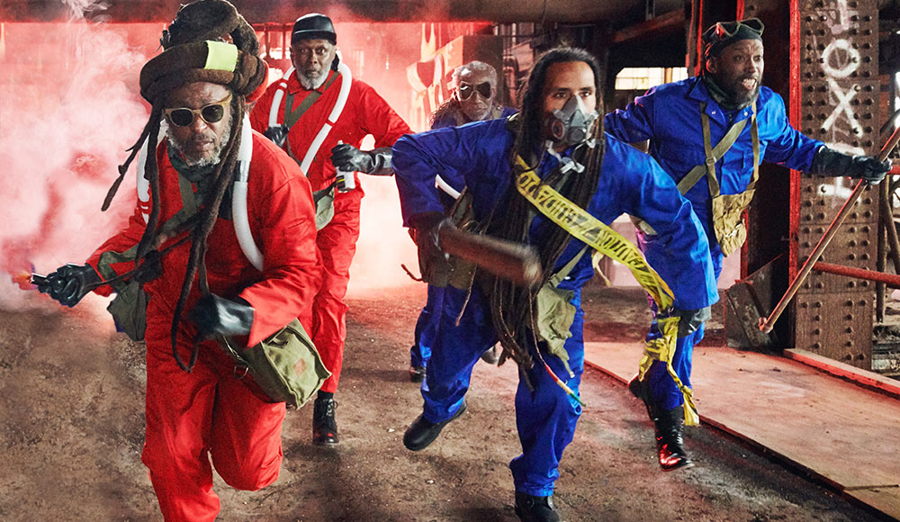 Four musicians leaning forward, grouped together, wearing jumpsuits (some bright red, some bright blue)