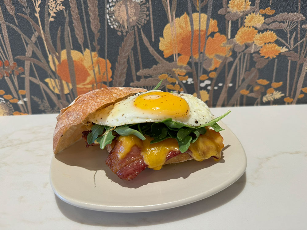 Bun with an egg, bacon and cheese inside sitting on a plate with a floral wallpapered wall behind it.