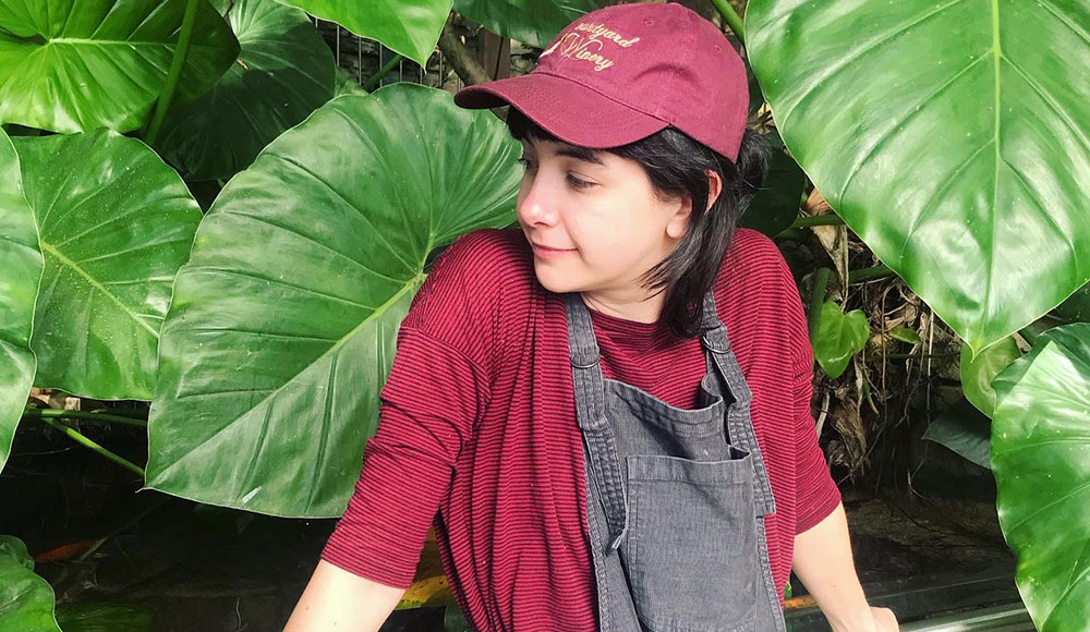Person dressed in red shirt and hat in overalls in front of a lush green plant