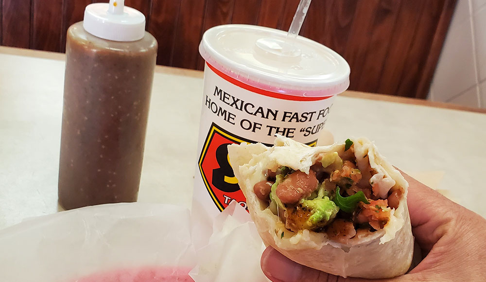 Hand holding a burrito with a drink and hot sauce dispenser also on the table