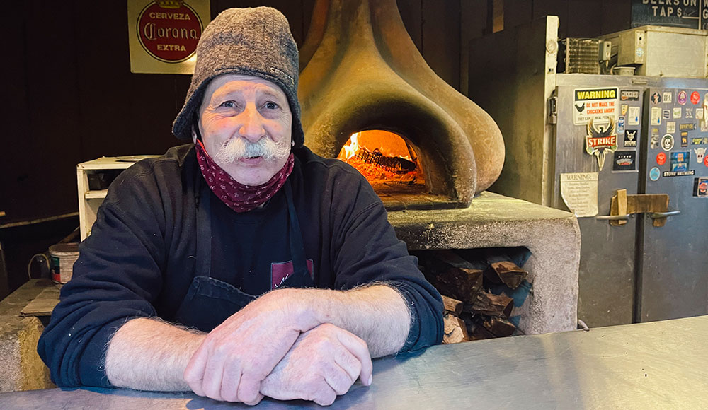 Man wearing a cap sitting in front of a wood-fired oven