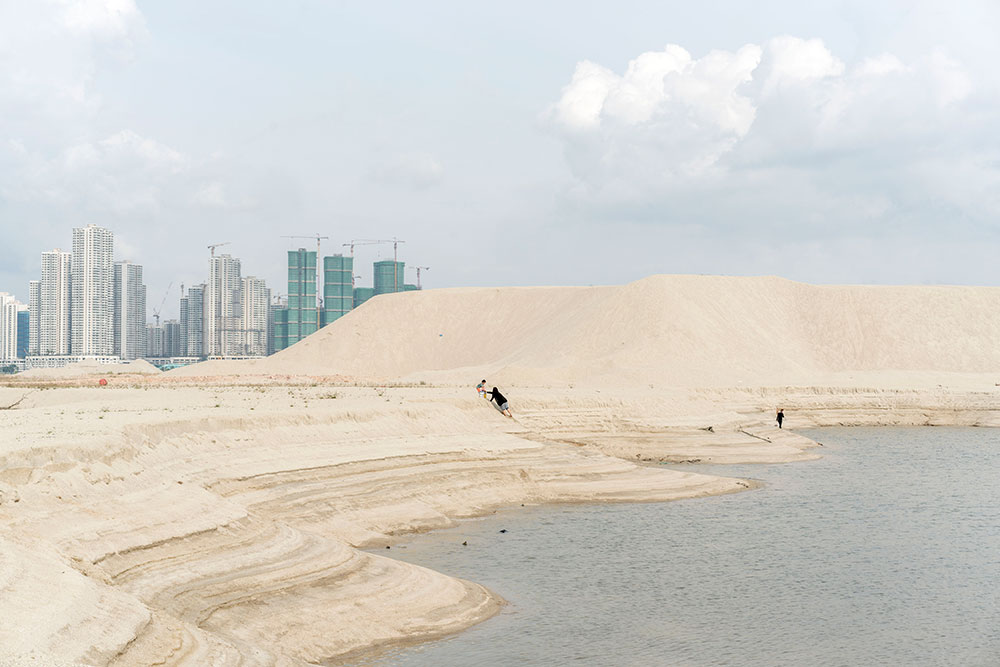 People climbing on sand dunes near water, with industrial buildings in the background