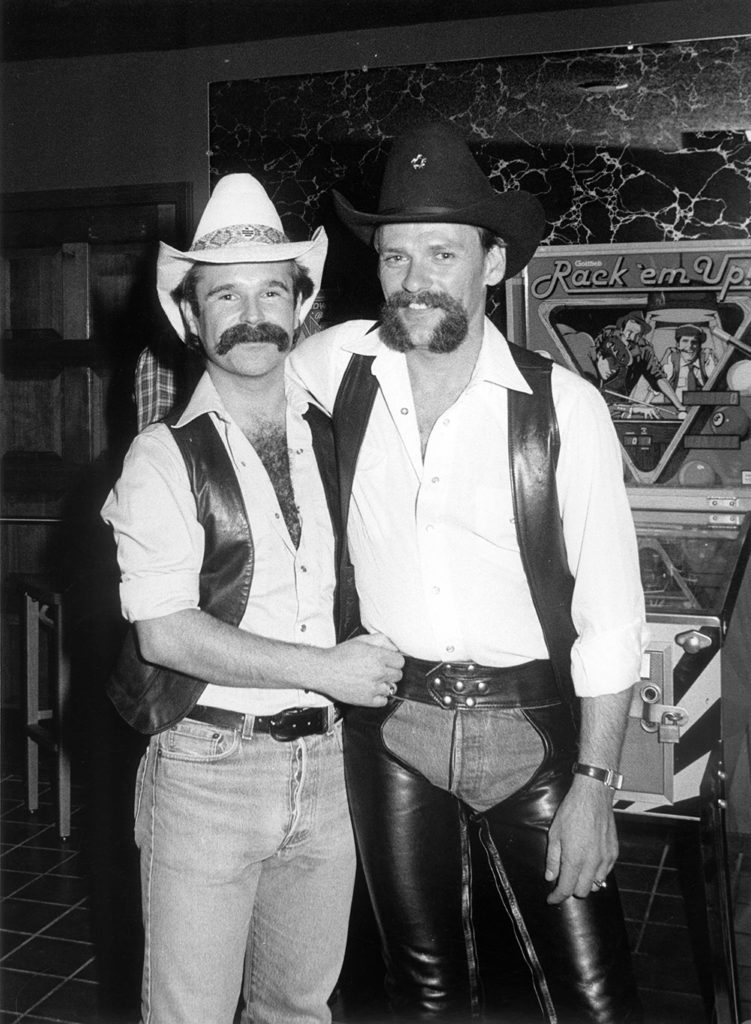 Two men dressed as cowboys, standing close together in a nightclub