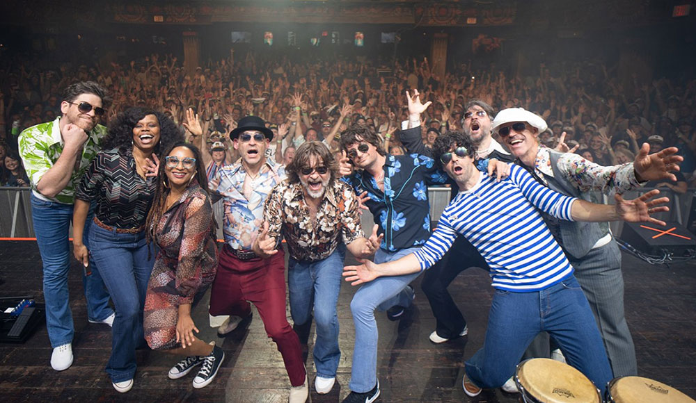 Large group of musicians posing for a shot on stage, with the audience seen behind them