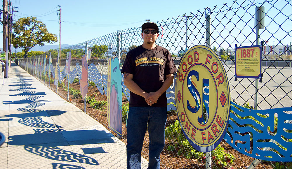 Man standing next to a chainlink fence with a piece of art hanging on i
