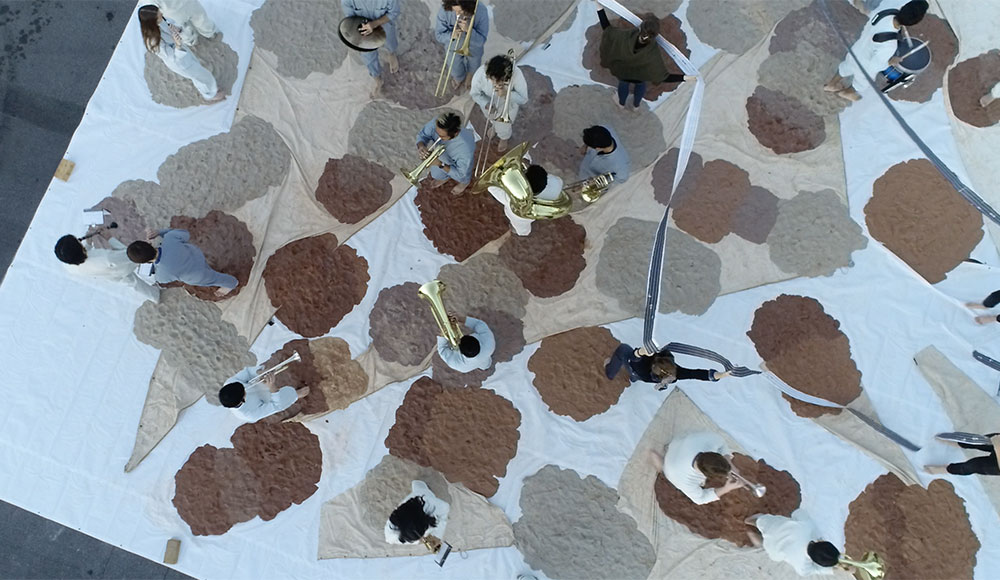 Overhead view of people playing instruments while walking on a white dropcloth with brown splotches of clay