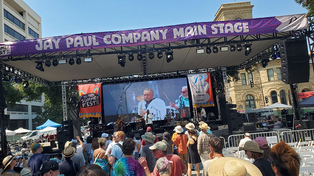 Musicians playing on an outdoor stage with a large screen offering a closeup of their faces