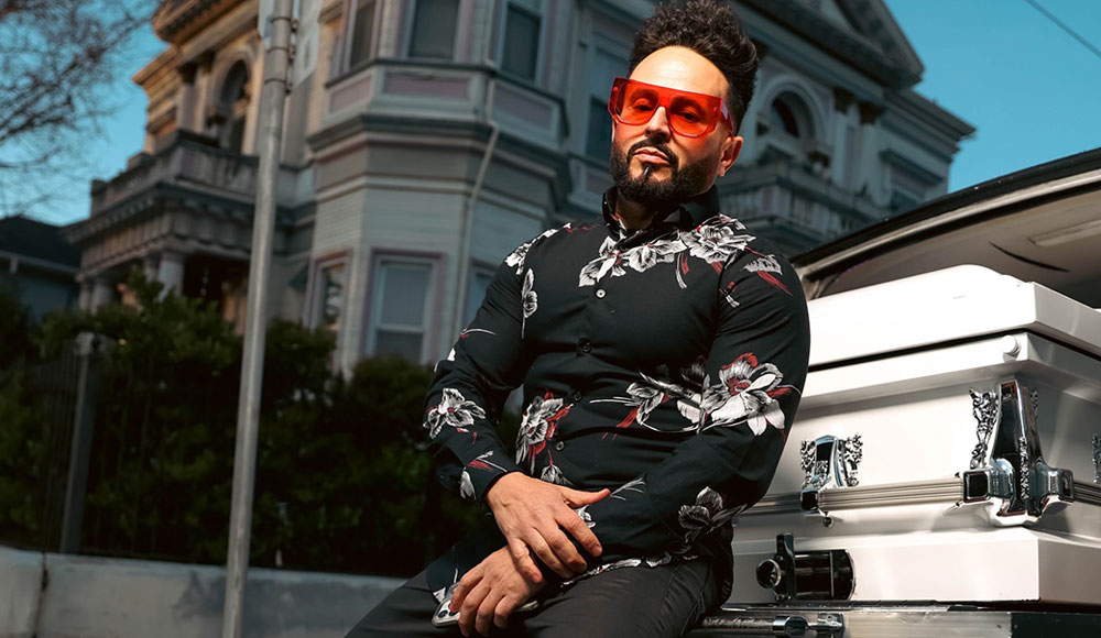 Man leaning against a casket in front of a Victorian house