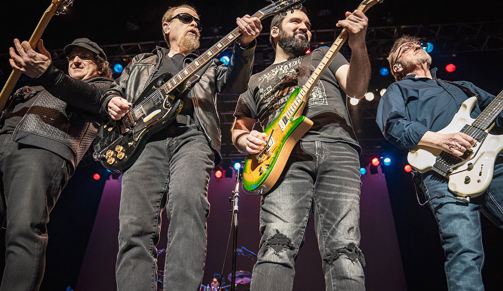Four men standing close together and playing guitars onstage