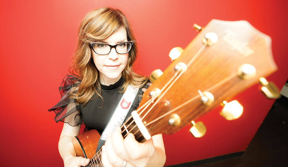 Woman playing a guitar against a red background, and her guitar is so close to the camera that it is out of focus