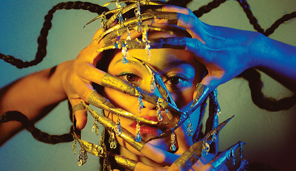 Woman with her hands in front of her face, painted blue and gold, with long decorative fingernails attached