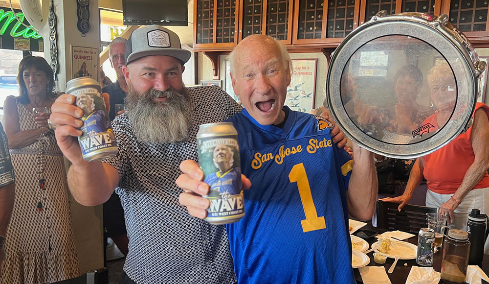 Two men holding up cans of beer, and one has a hand drum